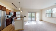 Located in Menomonee Falls, Wisconsin, this single-floor residence offers a well-designed open-concept layout that seamlessly connects the kitchen and living area. The kitchen is outfitted with rich dark-wood cabinetry, stainless steel appliances including a refrigerator, range, and microwave, and light hardwood-style flooring. A peninsula with a sink and counter seating serves as a natural dividing point between the kitchen and the carpeted living space. Pendant lights and track lighting provide ample illumination throughout.

The living area is bright and spacious, benefiting from natural light that streams through large sliding glass doors opening to a private balcony with outdoor views of surrounding greenery. The bedrooms feature neutral carpet, generous closet space, and easy access to the hallway. An in-unit laundry area adds everyday convenience.

Two bathrooms serve the residence: one featuring a walk-in glass-enclosed shower, dark cabinetry, a double-sink vanity, and wood-look flooring; the other offering a tub-shower combination. The overall aesthetic is clean and contemporary, with warm neutral tones and practical finishes throughout.