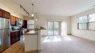 Located in Menomonee Falls, Wisconsin, this single-floor residence offers a well-designed open-concept layout that seamlessly connects the kitchen and living area. The kitchen is outfitted with rich dark-wood cabinetry, stainless steel appliances including a refrigerator, range, and microwave, and light hardwood-style flooring. A peninsula with a sink and counter seating serves as a natural dividing point between the kitchen and the carpeted living space. Pendant lights and track lighting provide ample illumination throughout.

The living area is bright and spacious, benefiting from natural light that streams through large sliding glass doors opening to a private balcony with outdoor views of surrounding greenery. The bedrooms feature neutral carpet, generous closet space, and easy access to the hallway. An in-unit laundry area adds everyday convenience.

Two bathrooms serve the residence: one featuring a walk-in glass-enclosed shower, dark cabinetry, a double-sink vanity, and wood-look flooring; the other offering a tub-shower combination. The overall aesthetic is clean and contemporary, with warm neutral tones and practical finishes throughout.