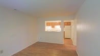 This single-floor space offers a straightforward, functional layout designed to maximize usability. The living area features light-toned hardwood flooring that extends throughout, contributing to a clean and cohesive aesthetic. Neutral white walls keep the space feeling bright and open, providing a versatile backdrop for a variety of interior styles.

A defining architectural feature is the pass-through window cut into the wall between the living area and the kitchen, creating a visual and functional connection between the two spaces. This design element enhances the sense of openness while maintaining a degree of separation. The kitchen is equipped with warm honey-toned wood cabinetry that lines the upper walls, a white refrigerator, and a sink positioned beneath under-cabinet lighting. The kitchen also benefits from a direct entryway accessible from the living area, allowing for easy circulation throughout the unit.

The overall ambiance is clean and understated, with a no-frills design that prioritizes practicality. The consistent flooring and neutral palette create a unified feel across the space, making it well-suited for those seeking a move-in-ready environment.
