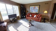 This single-floor residence features a thoughtfully arranged layout that maximizes its comfortable footprint across four distinct areas. The living room serves as the heart of the home, finished with plush carpeting and warm earth-toned walls that create an inviting atmosphere. A large window allows natural light to fill the space, complementing the cozy seating area centered around a glass-top coffee table. Bold wall art adds a decorative focal point to the room.

Adjacent to the living area, the dining space accommodates a glass-and-metal table with seating for four, positioned conveniently near the kitchen for easy entertaining. The kitchen is efficiently designed with white cabinetry, light countertops, a double sink, and tile flooring. Appliances including a refrigerator and range are seamlessly integrated into the layout.

The bedroom is a well-proportioned retreat featuring soft carpeting, a large window dressed with curtain panels that invite natural light, and ample space for a full furniture arrangement including a dresser and nightstands. Throughout the residence, the consistent warm brown palette and tile-to-carpet transitions create a unified, welcoming ambiance.