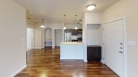 This single-floor residence is thoughtfully designed with a cohesive blend of functional and stylish elements throughout. The galley-style kitchen is a standout feature, showcasing rich, dark espresso cabinetry with raised-panel detailing that extends from base to upper cabinets, complemented by light granite-look countertops. Stainless steel appliances, including a refrigerator, microwave, and range, are paired with a double stainless sink and track lighting overhead, creating a well-equipped cooking space. A kitchen island with pendant lighting connects the kitchen to the open living area, which features warm hardwood-style flooring that runs throughout the main living spaces. The living area is generously proportioned, offering a ceiling fan, natural light from a window, and access to an exterior door. An arched niche adds an architectural detail to the living space. Bedrooms are finished with plush carpet and natural light from windows with blinds. The bathroom includes a soaking tub with a shower combo, tile flooring, a granite-look vanity countertop, and a natural light source from a window above the tub.