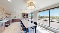 Located in Northglenn, Colorado, this single-floor studio and kitchen space offers a thoughtfully designed environment well-suited for creative workshops and group activities. The open layout maximizes functionality, seamlessly blending a dining and kitchen area with a dedicated workspace.

The studio area is defined by a long, central work table lined with contemporary black chairs, providing ample seating for group sessions. Large picture windows flood the room with natural light and offer open views of the surrounding neighborhood and Colorado's blue skies, creating a bright and inviting atmosphere. Elegant drum pendant lights hang overhead, complementing the light-toned tile flooring that runs throughout the space.

The kitchen area is a standout feature, showcasing rich walnut cabinetry with dark hardware, generous grey countertops, and a striking textured brick-style tile accent wall that stretches the full length of the room. Floating wooden shelves add both style and practical storage above the countertops. Recessed ceiling lighting enhances the warm tones of the cabinetry and tile, creating a welcoming ambiance.

Together, the two zones form a cohesive, modern space that balances practicality with aesthetic appeal, making it ideal for art classes, community gatherings, or creative workshops.