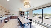 Located in Northglenn, Colorado, this single-floor studio and kitchen space offers a thoughtfully designed environment well-suited for creative workshops and group activities. The open layout maximizes functionality, seamlessly blending a dining and kitchen area with a dedicated workspace.

The studio area is defined by a long, central work table lined with contemporary black chairs, providing ample seating for group sessions. Large picture windows flood the room with natural light and offer open views of the surrounding neighborhood and Colorado's blue skies, creating a bright and inviting atmosphere. Elegant drum pendant lights hang overhead, complementing the light-toned tile flooring that runs throughout the space.

The kitchen area is a standout feature, showcasing rich walnut cabinetry with dark hardware, generous grey countertops, and a striking textured brick-style tile accent wall that stretches the full length of the room. Floating wooden shelves add both style and practical storage above the countertops. Recessed ceiling lighting enhances the warm tones of the cabinetry and tile, creating a welcoming ambiance.

Together, the two zones form a cohesive, modern space that balances practicality with aesthetic appeal, making it ideal for art classes, community gatherings, or creative workshops.