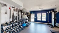 This fitness center is a thoughtfully designed exercise space that delivers a full gym experience within a compact, single-floor layout. The room is defined by bold navy blue accent walls paired with crisp white ceilings and trim, creating a dynamic and motivating atmosphere. Large windows and a glass door invite natural light throughout, offering outdoor views that enhance the open feel of the space. Ceiling fans ensure consistent airflow, contributing to a comfortable workout environment.

The strength training area is well-appointed, featuring a full dumbbell rack ranging across multiple weights, medicine balls, stability and exercise balls organized on a dedicated storage rack, a cable machine, and an adjustable workout bench. A large wall-mounted mirror spans one side of the room, allowing for form checks during exercises. A dramatic full-wall athletic mural adds an energizing visual focal point to the strength zone.

The cardio section houses a treadmill, elliptical trainer, stationary bike, and rowing machine, each positioned to take advantage of the natural light and outdoor views. Durable flooring designed for fitness use covers the entire space. Additional amenities include a sanitation station and posted fitness center rules, noting the space is available 24 hours. The overall design balances functionality and aesthetics to support a wide range of workout routines.
