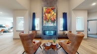 This single-floor community amenity space is thoughtfully designed to blend style with functionality. The main lounge area is an immediate focal point, boasting soaring ceilings, light hardwood flooring, and a dramatic floor-to-ceiling tile fireplace surround topped with a vibrant large-scale floral artwork. Flanking built-in window seats and rich navy drapes frame the natural light beautifully, while a pair of cognac leather wingback chairs and a live-edge wood coffee table create an inviting conversational arrangement. The open gathering area transitions seamlessly to a dining nook accented by a bold teal feature wall and a striking herringbone wood-panel accent wall, complemented by a rustic farmhouse table and eclectic mixed seating. Adjacent is a well-appointed kitchenette featuring granite countertops, dark cabinetry, stainless steel appliances, and a breakfast bar with seating — ideal for community events. The fitness center is generously sized, outfitted with strength and cable machines, a full dumbbell rack, treadmills, an elliptical, a spin bike, TRX suspension trainers, and wall-to-wall mirrors, all under ceiling fans and recessed lighting. Together, these spaces offer a comprehensive and design-forward amenity experience.