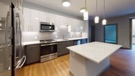 Located at 66 Fountain St in Providence, Rhode Island, this modern single-floor residence showcases a thoughtfully designed interior with clean lines and a neutral color palette throughout. The open-concept kitchen is a standout feature, equipped with stainless steel appliances, two-toned cabinetry in white and gray, a hexagonal tile backsplash, and a central island topped with a striking marble-look quartz countertop. Pendant lighting adds a refined touch to the space. Warm hardwood-style flooring flows through the kitchen and living area, where sliding glass doors lead to a private balcony with views of the surrounding neighborhood. The bedrooms are finished with soft carpeting and feature generous natural light from well-positioned windows. Each bedroom includes walk-in closet space outfitted with wire shelving systems for organized storage. The residence includes two full bathrooms: one with a sleek glass-enclosed walk-in shower featuring white subway tile, and a second with a full soaking tub and matching subway tile surround. Both bathrooms are finished with large-format floor tile, dark wood-toned vanities, white countertops, and expansive mirrors. The overall ambiance is clean, contemporary, and move-in ready.