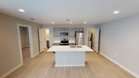 This single-floor space showcases a contemporary open-concept layout centered around a well-appointed kitchen. The kitchen serves as the focal point of the space, featuring a large white central island with an integrated sink, providing both a functional workspace and a natural gathering point. Stainless steel appliances, including a French door refrigerator, electric range, and built-in microwave, complement the gray cabinetry and white quartz-style countertops. A subtle tile backsplash adds texture behind the range, while recessed lighting overhead ensures consistent, even illumination throughout the area.

Light hardwood-style flooring extends across the open floor plan, contributing to a clean and cohesive aesthetic. The neutral gray walls and white trim create a bright, airy atmosphere. Multiple doorways visible from the main area lead to additional rooms and what appears to be a bathroom, offering a functional layout with clear sightlines throughout. The overall design is modern and minimalist, emphasizing clean lines, neutral tones, and practical finishes that balance style with everyday usability.