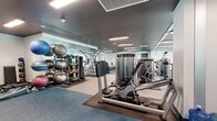 Located at McAnsh Square in Sarasota, FL, this contemporary fitness center offers a spacious, single-floor layout designed to accommodate a comprehensive range of workout activities. The facility is thoughtfully organized into distinct zones, maximizing functionality and user flow throughout the space.

The cardio zone is positioned along a wall of expansive floor-to-ceiling windows, allowing abundant natural light to pour in and providing views of the surrounding area. Life Fitness treadmills and elliptical machines are neatly lined up here, accompanied by vibrant teal accent walls adorned with geometric artwork that energizes the atmosphere.

The strength training area features a robust selection of selectorized cable and resistance machines, along with a dedicated free weights section housing a full rack of dumbbells, adjustable benches, and stability balls. Large wall-mounted mirrors throughout this zone enhance the sense of space and allow users to monitor their form.

Rubber flooring underlays are strategically placed beneath equipment, while blue commercial carpet covers the remaining areas, contributing to a clean, professional aesthetic. The facility also includes a water cooler station, towel service area, and wall-mounted television. Recessed and linear LED lighting ensures the entire space is well-illuminated. The overall design is modern and functional, providing an inviting environment for fitness pursuits in downtown Sarasota.
