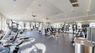 This fitness center offers a generously sized, single-floor layout designed to accommodate a comprehensive range of workout activities. The space is defined by its elegant architectural details, including a series of arched windows along the perimeter walls that allow abundant natural light to fill the room and provide views of the surrounding exterior. A two-toned wall treatment featuring a bold navy blue accent band adds a polished, purposeful aesthetic to the interior.

The ceiling features crown molding and is fitted with multiple ceiling fans, ensuring consistent air circulation throughout the space. Recessed lighting supplements natural light, maintaining a bright and energizing atmosphere. Full-wall mirrors line one side of the room, visually expanding the space while allowing users to monitor their form during exercise.

The flooring consists of durable, dark rubberized material, appropriate for heavy equipment and high-traffic use. The equipment layout is thoughtfully organized, with dedicated zones for strength training — including cable machines, free-weight racks, plate-loaded bars, and adjustable benches — as well as a cardio section featuring treadmills, stationary bikes, ellipticals, and stair climbers. The overall ambiance is that of a well-maintained, professional-grade fitness facility.