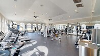 This fitness center offers a generously sized, single-floor layout designed to accommodate a comprehensive range of workout activities. The space is defined by its elegant architectural details, including a series of arched windows along the perimeter walls that allow abundant natural light to fill the room and provide views of the surrounding exterior. A two-toned wall treatment featuring a bold navy blue accent band adds a polished, purposeful aesthetic to the interior.

The ceiling features crown molding and is fitted with multiple ceiling fans, ensuring consistent air circulation throughout the space. Recessed lighting supplements natural light, maintaining a bright and energizing atmosphere. Full-wall mirrors line one side of the room, visually expanding the space while allowing users to monitor their form during exercise.

The flooring consists of durable, dark rubberized material, appropriate for heavy equipment and high-traffic use. The equipment layout is thoughtfully organized, with dedicated zones for strength training — including cable machines, free-weight racks, plate-loaded bars, and adjustable benches — as well as a cardio section featuring treadmills, stationary bikes, ellipticals, and stair climbers. The overall ambiance is that of a well-maintained, professional-grade fitness facility.