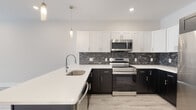 This well-appointed single-floor space showcases a thoughtfully designed, contemporary interior with a clean, neutral palette and quality finishes throughout. The kitchen is a standout feature, combining crisp white upper cabinetry with deep espresso-toned lower cabinets, all set against a striking dark marble-patterned tile backsplash. White quartz countertops extend across the workspace and peninsula, providing ample prep and serving surface. A full suite of stainless steel appliances — including a French door refrigerator, electric range, over-the-range microwave, and dishwasher — completes the kitchen. Pendant lighting above the peninsula adds a modern accent. Light-toned hardwood-style flooring flows seamlessly from the kitchen into the open adjoining living area, which benefits from natural light through large windows offering leafy outdoor views. The bathroom is finished with large-format grey stone-look wall tiles surrounding a soaking tub and shower combination, complemented by dark marble-effect floor tiles. A floating wood-toned vanity with a white basin and mirrored medicine cabinet adds a refined, modern touch. Throughout the space, recessed lighting, white walls, and clean architectural lines create a bright, welcoming ambiance.