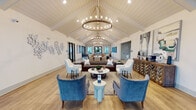 This single-story leasing office in Destin, Florida, is designed to make a striking first impression through thoughtful coastal-inspired interiors and a well-organized layout encompassing a lobby lounge, seating areas, and a game room. The centerpiece of the space is the expansive lobby lounge, defined by soaring vaulted ceilings with white-painted shiplap detailing and exposed wood beams. A large circular chandelier with exposed Edison bulbs anchors the room, casting warm light over multiple seating arrangements that include plush blue velvet armchairs, a rich brown leather sofa, and white accent chairs, all positioned atop patterned area rugs over light wood-style flooring. Large windows and glass doors flood the interior with natural light and offer views of the surrounding landscaping and outdoor deck area. A separate lounge nook features floor-to-ceiling blue shiplap walls, built-in shelving, a wall-mounted television, and curated décor, creating an intimate retreat within the broader space. Nearby, a functional kitchenette with sage green cabinetry, dark countertops, and an oversized woven pendant light serves as both a refreshment station and workspace. Throughout, bold patterned drapery, curated artwork, and carefully selected furnishings contribute to a polished, coastal-modern aesthetic.