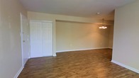 This single-floor space presents a straightforward, functional layout designed for comfortable living. The main living area is finished with rich, warm-toned hardwood-style flooring that extends throughout, providing a cohesive and inviting foundation. Neutral beige walls create a versatile backdrop, easily adaptable to a variety of interior styles and furnishing choices.

A notable feature of this space is the set of white double sliding closet doors, offering generous built-in storage that is both accessible and visually unobtrusive. The doors feature a classic paneled design that complements the clean, simple aesthetic of the room.

Overhead, a traditional multi-arm chandelier with warm-toned bulbs serves as a central lighting fixture, adding a subtle decorative touch to the dining or living area. The ceiling is smooth and consistent in height, contributing to the open feel of the space. White baseboards neatly frame the perimeter of the room, providing a crisp contrast against the flooring and walls.

Overall, the space maintains a clean, move-in-ready condition with a neutral palette, making it a practical and adaptable environment.