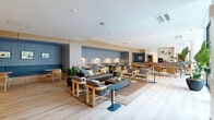 Located at 700 Hamilton Ave in Nashville, TN, this thoughtfully designed single-floor amenity and coworking space offers a generous, open layout across nearly 2,000 square feet. The interior showcases a contemporary aesthetic defined by light wood flooring, soft neutral tones, and blue textured wall panels that lend warmth and visual depth throughout.

The main open area is organized into distinct functional zones, including a relaxed lounge with a channel-tufted leather sofa, rounded upholstered armchairs, and a low stone-topped coffee table — ideal for informal collaboration or quiet relaxation. Adjacent to this is a dedicated coworking section featuring individual semi-private booths with built-in task lighting set against blue fabric wall panels, alongside communal worktables with ergonomic seating and personal dividers.

A sunlit café-style seating area lines a floor-to-ceiling glass curtain wall, offering direct views of a landscaped courtyard and ample natural light. The space also includes a private conference room with a round wood table, wainscoted walls, and warm gold accents, as well as a secondary work room with a coffee station and built-in shelving. Lush potted greenery is integrated throughout, enhancing the fresh, inviting ambiance of this well-appointed Nashville workspace.