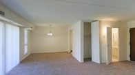 This two-floor space is designed with simplicity and functionality in mind. The interior showcases neutral cream-toned walls throughout, complemented by wall-to-wall carpeting in a warm beige hue that adds a sense of comfort underfoot. The open layout of the main living area allows for versatile furniture arrangement, making the space feel welcoming and adaptable.

A small chandelier with multiple candle-style lights serves as a central lighting fixture, adding a modest decorative touch to the room. The ceiling is smooth and painted white, contributing to the overall brightness of the interior. Visible HVAC venting is integrated into the ceiling, ensuring climate control throughout.

A notable feature of this space is the built-in closet, positioned along a partial wall divider. The closet includes a hanging rod and built-in shelving, offering practical storage solutions. The bifold-style white door complements the clean, neutral aesthetic of the surrounding walls.

A doorway visible in the background leads to an adjacent room or hallway, suggesting a connected and thoughtfully arranged floor plan. Overall, the space maintains a clean, understated character suitable for a variety of uses.