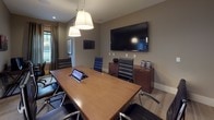 This conference room offers a well-appointed, functional setting designed to support productive meetings and collaborative work. The centerpiece of the space is a substantial wood conference table surrounded by black leather executive chairs, providing comfortable seating for multiple attendees. Overhead, a pair of pendant drum lights suspended from a track system cast warm, focused illumination across the table surface, complementing the recessed ceiling lighting throughout.

A large wall-mounted flat-screen display serves as the primary presentation focal point, positioned for clear sightlines from all seating positions. A built-in credenza beneath the screen provides additional storage, while a tablet integrated into the table surface supports connectivity and meeting management. A dedicated workstation with a desktop computer is situated along one wall, adding a secondary workspace within the room.

The space is finished with neutral taupe walls, light carpeting, and crisp white baseboards, creating a clean and professional atmosphere. A full-height glass partition with a sliding panel opens the room to an adjacent bright common area, allowing natural light to filter in while maintaining the option for privacy. Framed artwork and decorative accents contribute a polished, refined character to the overall environment.