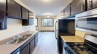 This single-floor residential unit offers a practical and functional layout designed for comfortable everyday living. The galley-style kitchen is outfitted with dark espresso cabinetry lining both walls, light wood-look flooring, a double-basin sink, laminate countertops, and black appliances including a refrigerator and gas range. A window at the far end of the kitchen brings in natural light and overlooks surrounding trees, creating a pleasant connection to the outdoors.

The living area is generously proportioned, featuring neutral carpet, cream-toned walls with dark baseboards, and a wall-mounted air conditioning unit alongside baseboard heating for year-round climate control. A large window frames views of the natural landscape outside.

The bedroom is well-sized and carpeted, with a ceiling fan, a large set of dark wood bi-fold closet doors offering ample storage, and a secondary door providing easy access to the rest of the unit. Baseboard heating is present here as well.

The bathroom features a full-size soaking tub and shower combo with clean white surround walls, a toilet, and warm neutral wall tones. Throughout the unit, dark wood trim and door accents provide visual contrast against the light walls and carpet, contributing to a cohesive and understated aesthetic.