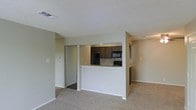 This single-floor residence offers a practical and well-organized layout designed for comfortable everyday living. The open-concept living area is finished with wall-to-wall carpet, creating a warm and inviting atmosphere throughout the main space. A textured ceiling adds subtle dimension overhead, while neutral-toned walls provide a clean, versatile backdrop.

The kitchen is partially enclosed by a half-wall peninsula that serves as both a visual divider and a functional breakfast bar, topped with a light granite-style countertop. Dark espresso-toned cabinetry lines the kitchen walls, offering ample storage, while the space is equipped with black appliances including a refrigerator, microwave, and range. Under-cabinet lighting adds a practical touch to the cooking area.

A doorway visible from the main living area leads to additional rooms, suggesting a thoughtful and connected floor plan. The space is served by central air and heating, with ceiling vents positioned throughout for consistent climate control. Clean transitions between the carpeted living zone and the hard-floored kitchen area help define each functional space clearly. Overall, this residence presents a straightforward and functional living environment with essential amenities well-integrated into its compact, single-level design.