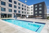 This outdoor amenity deck in Boise, Idaho offers a thoughtfully designed resort-style experience set against a backdrop of modern multi-story architecture. At the heart of the space is a generously sized rectangular swimming pool with crystal-clear turquoise water, accessible via wide entry steps with stainless steel railings. The surrounding pool deck is finished in large-format concrete pavers, providing ample space for an extensive arrangement of lounge chairs and umbrellas, ideal for sunbathing under Boise's abundant sunshine. Decorative river rock landscaping beds add a natural, textural contrast to the clean hardscape surfaces, while horizontal wood-slat privacy screens define portions of the perimeter with a warm aesthetic detail. Beyond the pool, a spacious courtyard area features multiple seating arrangements, outdoor grilling stations, and a fire pit lounge—perfect for social gatherings. A patch of artificial turf accommodates lawn games, adding a recreational element to the deck. The space benefits from open sky exposure and sweeping views of the Boise city skyline and surrounding mountain foothills. The overall design balances contemporary urban style with comfortable, functional outdoor living, making it a standout amenity within its Boise community.