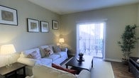 This single-floor space centers around a well-appointed living room designed for comfort and functionality. The room is finished in soft, neutral grey-green tones that create a calm and inviting atmosphere. White slipcovered sofas arranged around a dark wood coffee table provide ample seating, while decorative throw pillows in warm earth tones add visual interest and a welcoming feel.

A standout feature of the living room is the full-height sliding glass door fitted with vertical blinds, which floods the space with natural light and provides direct access to an outdoor balcony. The balcony is visible through the glass and appears to include seating, extending the usable living area outdoors.

Warm ambient lighting from a classic table lamp complements the natural light, enhancing the room's cozy character during evening hours. The walls are accented with a small collection of framed artwork, adding a tasteful decorative touch. Light-toned carpeting runs throughout the living area, contributing to the overall softness of the interior. The space presents a practical yet comfortable layout suited for everyday living.