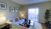This single-floor space centers around a well-appointed living room designed for comfort and functionality. The room is finished in soft, neutral grey-green tones that create a calm and inviting atmosphere. White slipcovered sofas arranged around a dark wood coffee table provide ample seating, while decorative throw pillows in warm earth tones add visual interest and a welcoming feel.

A standout feature of the living room is the full-height sliding glass door fitted with vertical blinds, which floods the space with natural light and provides direct access to an outdoor balcony. The balcony is visible through the glass and appears to include seating, extending the usable living area outdoors.

Warm ambient lighting from a classic table lamp complements the natural light, enhancing the room's cozy character during evening hours. The walls are accented with a small collection of framed artwork, adding a tasteful decorative touch. Light-toned carpeting runs throughout the living area, contributing to the overall softness of the interior. The space presents a practical yet comfortable layout suited for everyday living.