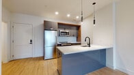 Located at 20 Grand in Englewood, NJ, this thoughtfully designed single-floor residence combines modern aesthetics with functional living. The open-concept kitchen serves as the centerpiece, showcasing sleek gray cabinetry accented by warm wood trim, a light blue glass island base topped with a striking white quartz countertop, and stainless steel appliances including a refrigerator and microwave. A matte black faucet and light blue glass tile backsplash add refined finishing touches, while cylindrical pendant lights hang above the island, casting a warm, focused glow. Light hardwood flooring flows consistently throughout the unit, complementing the bright, neutral-toned walls and creating an airy, cohesive atmosphere. The living area offers generous proportions with direct access to a private balcony through a glass-panel door, inviting natural light and outdoor views into the space. The bedroom is well-proportioned and features its own window with exterior views. The bathroom is finished with a floating wood vanity, an illuminated mirror, large-format marble-style wall tiles surrounding the bathtub, and matte black fixtures throughout. A dedicated laundry closet with stacked washer and dryer units adds everyday convenience to this polished, move-in-ready residence.