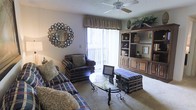 This single-floor space centers around a comfortable living room designed for both relaxation and functionality. The room is anchored by a large wooden entertainment unit with open shelving and enclosed cabinetry, providing ample display and storage space. Plaid-upholstered seating, including a sofa and armchair, gives the room a classic, homey character, complemented by a glass-topped coffee table with decorative ironwork legs and a matching plaid ottoman. A ceiling fan overhead provides both air circulation and ambient lighting. Natural light flows into the room through a sliding glass door dressed with vertical blinds and a fabric valance, offering access to an outdoor balcony or patio. Carpeted flooring adds warmth underfoot, while a decorative round mirror with an ornate frame serves as a focal wall accent. The textured ceiling adds a traditional architectural finish. Overall, the space presents a warm, inviting ambiance with a well-coordinated traditional aesthetic, making it well-suited for everyday living and entertaining.