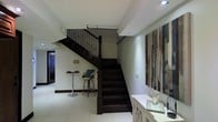 This two-storey space makes a strong first impression through its central staircase, crafted from dark wood with ornamental wrought-iron balusters and a sturdy newel post. The staircase serves as a visual focal point, drawing the eye upward and connecting both levels with both function and style. The ground floor features light-toned tile flooring that contrasts effectively with the dark wood elements, lending the space a clean, balanced aesthetic. Recessed ceiling lighting provides a warm, focused glow throughout the interior, highlighting key areas including a console table along one wall and a compact bistro-style bar setup with a round table and cushioned stool nearby. A large, multi-panel abstract artwork adorns one wall, adding texture and depth to the otherwise neutral color palette of whites and creams. The lower level also features a corridor leading to additional rooms, suggesting a well-organized layout. The overall ambiance is polished and contemporary, combining classic architectural details with modern decorative touches to create a refined and comfortable living environment across both floors.