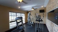 This single-floor fitness center is designed to provide a comprehensive workout environment with a practical and functional layout. The space is outfitted with a variety of exercise equipment, including a treadmill, an elliptical trainer, and a multi-function cable machine, catering to both cardio and strength training needs. A reference chart is conveniently mounted on the wall near the equipment, offering exercise guidance for users. A wall-mounted flat-screen television adds an entertainment element to workout sessions. The room features warm yellow-toned walls that create an inviting atmosphere, complemented by dark rubber flooring that is both durable and slip-resistant — ideal for a fitness setting. Two ceiling fans help maintain air circulation throughout the space, ensuring comfort during intense workouts. Large windows fitted with slatted blinds line one wall, allowing ample natural light to filter in while offering adjustable privacy. Recessed lighting in the ceiling provides additional illumination. The overall ambiance is welcoming and well-maintained, making it a practical and comfortable space for a full range of fitness activities.