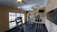 This single-floor fitness center is designed to provide a comprehensive workout environment with a practical and functional layout. The space is outfitted with a variety of exercise equipment, including a treadmill, an elliptical trainer, and a multi-function cable machine, catering to both cardio and strength training needs. A reference chart is conveniently mounted on the wall near the equipment, offering exercise guidance for users. A wall-mounted flat-screen television adds an entertainment element to workout sessions. The room features warm yellow-toned walls that create an inviting atmosphere, complemented by dark rubber flooring that is both durable and slip-resistant — ideal for a fitness setting. Two ceiling fans help maintain air circulation throughout the space, ensuring comfort during intense workouts. Large windows fitted with slatted blinds line one wall, allowing ample natural light to filter in while offering adjustable privacy. Recessed lighting in the ceiling provides additional illumination. The overall ambiance is welcoming and well-maintained, making it a practical and comfortable space for a full range of fitness activities.