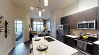 This single-floor residence showcases an open-concept design that seamlessly connects the kitchen and living areas into one cohesive, functional space. The kitchen is a standout feature, equipped with sleek dark espresso cabinetry that contrasts elegantly against bright white quartz countertops. A large kitchen island serves as both a prep space and a casual dining area, outfitted with an undermount sink and pendant lighting overhead. Stainless steel appliances, including a French door refrigerator and range, add a polished, modern touch. A neutral tile backsplash completes the refined aesthetic. The living area beyond is bright and welcoming, benefiting from expansive windows that draw in generous natural light and offer views of the surroundings. A ceiling fan enhances comfort throughout the open space. Rich hardwood-style flooring runs continuously through both areas, grounding the design with warmth. Elevated ceilings and recessed track lighting contribute to an airy, spacious ambiance. The overall aesthetic balances contemporary style with functional comfort, making this a well-appointed and visually cohesive living environment.
