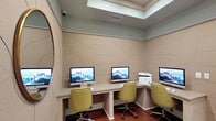 This single-floor business center is thoughtfully designed to offer a functional yet visually refined workspace. The room is lined with built-in, light wood desks arranged along the walls, accommodating multiple iMac workstations and a shared HP multifunction printer — providing everything needed for everyday computing and printing tasks. Plush, chartreuse upholstered rolling chairs with nailhead trim add a touch of sophisticated style while ensuring comfort during extended use. The walls are clad in textured, neutral-toned acoustic panels featuring subtle curved relief patterns, contributing both to the room's aesthetic warmth and its sound-dampening qualities. A tray ceiling with a softly lit skylight panel draws the eye upward, lending the space an open, airy feel. A round gold-framed mirror on one wall adds depth and a decorative focal point, while a pair of bold abstract artworks — one with cool tones and one with warm — brings vibrant artistic energy to the entryway wall. Rich terracotta-hued tile flooring grounds the space with warm contrast. The center opens to an adjacent common area, suggesting seamless connectivity to broader amenities. Overall, the space balances practicality with contemporary interior design.