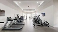 Located in Fort Myers, FL, this well-appointed fitness center offers a comprehensive exercise environment designed to support a wide range of workout routines. The space features a clean, modern aesthetic with crisp white walls, a tray ceiling with recessed lighting, and durable rubber flooring throughout, creating a professional and motivating atmosphere.

The cardio zone is generously equipped with multiple treadmills, elliptical trainers, stair climbers, and stationary bikes from brands including Matrix, providing users with a full selection of aerobic options. A wall-mounted flat-screen TV adds entertainment during workouts. Large windows and glass-panel doors along one wall allow ample natural light to fill the space while offering views of the Florida outdoors, including glimpses of palm trees.

The strength training area features a cable crossover machine, selectorized weight machines, and a rack of dumbbells, accommodating both machine-based and free-weight training. Adjustable Matrix benches are positioned for versatile use. A functional training station includes an assortment of medicine balls, stability balls of varying sizes, and resistance training accessories neatly organized on wall-mounted racks.

Large mirrors on one wall enhance the sense of space while allowing users to monitor their form. The facility also includes a water station and safety equipment, reflecting a commitment to member well-being. Ceiling fans contribute to comfort in the Florida climate.
