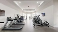 Located in Fort Myers, FL, this well-appointed fitness center offers a comprehensive exercise environment designed to support a wide range of workout routines. The space features a clean, modern aesthetic with crisp white walls, a tray ceiling with recessed lighting, and durable rubber flooring throughout, creating a professional and motivating atmosphere.

The cardio zone is generously equipped with multiple treadmills, elliptical trainers, stair climbers, and stationary bikes from brands including Matrix, providing users with a full selection of aerobic options. A wall-mounted flat-screen TV adds entertainment during workouts. Large windows and glass-panel doors along one wall allow ample natural light to fill the space while offering views of the Florida outdoors, including glimpses of palm trees.

The strength training area features a cable crossover machine, selectorized weight machines, and a rack of dumbbells, accommodating both machine-based and free-weight training. Adjustable Matrix benches are positioned for versatile use. A functional training station includes an assortment of medicine balls, stability balls of varying sizes, and resistance training accessories neatly organized on wall-mounted racks.

Large mirrors on one wall enhance the sense of space while allowing users to monitor their form. The facility also includes a water station and safety equipment, reflecting a commitment to member well-being. Ceiling fans contribute to comfort in the Florida climate.