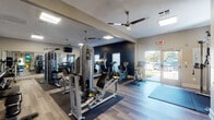 This single-floor fitness center is thoughtfully designed to support a full range of workout routines. The space features light wood-style flooring throughout, providing a clean and modern aesthetic that is both durable and visually appealing. Ceiling fans are installed across the facility to maintain comfortable airflow during workouts, complemented by recessed flush lighting that ensures the space is well-lit at all times.

The cardio zone is equipped with multiple treadmills, upright stationary bikes, and cycling/spin bikes, all positioned to take advantage of the natural light streaming through large windows and a glass-panel exterior door that opens to the outdoors. A bold navy accent wall anchors this section of the room, adding a dynamic visual contrast to the neutral tones throughout the rest of the space.

The strength training area offers a variety of cable and weight machines, a weight bench, and a dumbbell rack, catering to users of all fitness levels. Full-length mirrors line portions of the walls, enhancing the sense of openness and allowing users to monitor their form.

A distinctive glass partition wall separates the fitness center from an adjacent lounge or common area, maintaining visual connectivity between the spaces while providing a defined workout environment. Wall-mounted televisions are positioned throughout for entertainment during exercise sessions.
