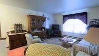 This single-floor residence offers a comfortably arranged and traditionally styled living space. The living room serves as the heart of the home, showcasing a prominent dark wood china hutch with glass-paneled doors, providing both storage and a classic decorative focal point. Soft, neutral-toned carpeting runs underfoot, contributing to the warm and inviting ambiance of the room. Seating is arranged thoughtfully around a glass-top coffee table, creating an intimate and functional gathering area. A flat-screen television is positioned on a dark wood media stand, complemented by a lush indoor plant that adds a touch of greenery to the interior. Natural light filters into the room through a window dressed with layered sheer curtains and rich, dark valances, offering a soft glow while maintaining a sense of privacy. A baseboard heating unit along the exterior wall suggests a practical and efficient approach to climate comfort. The overall aesthetic is one of classic, homey warmth — a space clearly designed with comfort and everyday livability in mind.