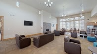 This single-floor community clubhouse is thoughtfully designed to offer a variety of shared amenities in a warm, inviting setting. The centerpiece of the space is a generously sized lounge area with soaring vaulted ceilings, elegant chandelier lighting, and a striking floor-to-ceiling stacked-stone fireplace. Dark leather sofas and armchairs are arranged throughout, creating comfortable seating for gathering or relaxing, while a wall-mounted flat-screen TV adds a modern touch. Carpet tile flooring in neutral tones grounds the space with a clean, contemporary feel.

Adjacent to the lounge is a bright dining area featuring rich hardwood flooring, tall windows dressed with warm wood-trimmed blinds that invite abundant natural light, and high-top tables with bar-style seating. A kitchenette with knotty alder cabinetry, a mosaic tile backsplash, granite countertops, stainless steel appliances, and a breakfast bar completes the social gathering area.

The fitness center is well-appointed across two connected rooms, offering a comprehensive selection of cardio equipment including treadmills and ellipticals, as well as strength-training machines, free weights, medicine balls, stability balls, and a stationary bike. Rubber flooring, ceiling fans, and natural light from multiple windows make this an energizing space for exercise.