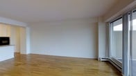 This single-floor space presents a clean, minimalist aesthetic that provides a welcoming blank canvas for a variety of uses. The interior is characterized by crisp white walls that reflect light throughout, contributing to an open and airy atmosphere. Warm-toned hardwood flooring runs across the main living area, adding a natural richness and a sense of continuity to the space. The layout is straightforward and efficient, with a generously proportioned main room that offers notable flexibility in how it can be arranged and utilized. A baseboard heating unit is visible along one wall, providing a practical climate-control solution integrated neatly into the design. Subtle architectural details, including clean wall transitions and a structured column element, add modest visual interest while maintaining the space's understated character. The neutral palette of whites and natural wood tones creates a calm, uncluttered ambiance that would suit a range of functions, from a residential living area to a professional or creative workspace. Overall, the space offers a simple yet refined environment with a strong foundation for customization.