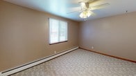 This single-floor space offers a straightforward, functional layout that is visible in its open floor plan. The living area is generously sized, featuring light-toned carpeting and a large window fitted with vertical blinds that allows natural light to pour in, casting bright streaks across the floor. A baseboard heating system runs along the walls, ensuring consistent warmth throughout the unit.

The bedrooms continue the carpeted aesthetic, each room outfitted with a ceiling fan and light fixture, horizontal window blinds, warm neutral-toned walls, and wood baseboard trim. Baseboard heaters are present in these rooms as well, contributing to a cohesive heating setup across the space.

The kitchen features warm oak cabinetry with open shelving, light laminate countertops, a tiled backsplash, and a distinctive double-basin green sink positioned beneath a window with horizontal blinds, providing natural light while cooking. Storage is ample with both upper and lower cabinets.

The bathroom is clean and functional, finished with light tile flooring and a tub-shower combination surrounded by white and accented square tiles. A window above the tub provides ventilation, and a wood-toned vanity with a white sink completes the space.