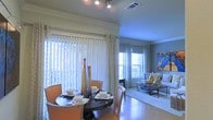 This single-floor residence seamlessly blends the dining and living areas into one cohesive, open-plan layout. The dining space is anchored by a dark, round table surrounded by upholstered chairs in warm cognac tones with nailhead trim detailing, lending a refined yet inviting character to the room. Decorative glass vases in amber and cobalt hues serve as a stylish centerpiece on the table.

Floor-to-ceiling vertical blinds and sheer curtain panels line the expansive windows, allowing diffused natural light to fill the space while maintaining privacy. Track lighting mounted on the ceiling provides additional ambient and task illumination, complementing the natural light throughout the day.

The adjoining living area is visible beyond a subtle architectural division, featuring a comfortable sofa dressed with accent pillows, a glass-top coffee table, and a vibrant piece of wall art that adds a pop of color. A second set of windows with blinds brings consistent natural light into the living space as well. Light-toned flooring runs throughout, unifying both areas and contributing to the open, airy feel of the residence.