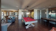 This thoughtfully designed single-floor amenity space blends rustic warmth with contemporary sophistication through a cohesive mix of reclaimed wood accent walls, exposed brick detailing, and rich dark hardwood flooring. The open layout is anchored by a striking game area showcasing a red-felt pool table beneath an elegant pendant light, creating an inviting focal point for social gatherings. Adjacent to the game area, a cozy lounge nook features built-in seating with warm ambient lighting, ideal for intimate conversation. A dedicated wet bar and kitchenette area is outfitted with sleek cabinetry, a herringbone tile backsplash, stainless steel appliances, and pendant lighting, complemented by a large oval dining table surrounded by leather and upholstered seating. The living lounge area is furnished with plush sofas and accent chairs arranged beneath a decorative drum chandelier, with large windows throughout allowing abundant natural light and outdoor views. A separate seating alcove with leather club chairs and a reclaimed wood accent wall offers a more private retreat. The space is further enhanced by curated artwork, layered area rugs, and architectural details that together create a refined yet approachable atmosphere suited for both relaxation and entertainment.