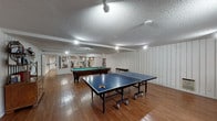 This single-floor space is designed for both recreation and fitness, offering a well-rounded amenity suite across several connected areas. The main game room is a generously sized, open area finished with warm hardwood flooring, white vertical wood-panel walls, and a textured ceiling with recessed and track lighting. It comfortably accommodates both a full-size pool table and a ping-pong table, making it an ideal gathering space for leisure and entertainment. A wooden cabinet provides additional storage along the wall.

Directly accessible from the game room is a dedicated fitness area, separated by a glass partition that allows natural sightlines between the two spaces. The gym is equipped with a treadmill, stationary bike, weight bench, and additional exercise equipment, all set on hardwood flooring. Full-length mirrors line the walls, enhancing the sense of space and functionality for workouts. Overhead fluorescent lighting ensures the area is well-lit throughout.

A connecting hallway with vertical white paneling and a floating glass shelf leads between the main areas. A relaxed lounge room features wicker chairs with blue cushions and large sliding glass doors that open to an outdoor patio with staircases, bringing in natural light and providing easy outdoor access. The cohesive white paneling throughout unifies the entire space with a clean, consistent aesthetic.