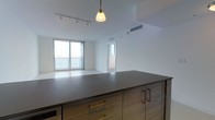 This contemporary single-floor space showcases a thoughtfully designed open-concept layout that seamlessly connects the kitchen and living areas. The kitchen is anchored by a substantial island finished in dark stone countertop with warm wood cabinetry and distinctive gold-toned hardware, adding a refined touch to the space. Overhead, a combination of a pendant light and a track lighting system provides both ambient and task illumination, complementing the clean white ceilings above.

The living area is bathed in natural light, courtesy of expansive floor-to-ceiling sliding glass windows and doors that frame breathtaking water views beyond. The open sightlines and bright, neutral tones throughout create an airy and inviting atmosphere. A sliding glass door leads to what appears to be a balcony, extending the living space outdoors and allowing for seamless indoor-outdoor connectivity.

Climate control is well-appointed, with visible HVAC vents integrated into the ceiling for year-round comfort. The overall aesthetic is clean, modern, and minimalist, with white walls and light flooring that enhance the sense of openness. The combination of contemporary finishes, panoramic views, and abundant natural light makes this a bright and sophisticated living space.