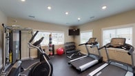 This dedicated fitness center is thoughtfully designed to support a wide range of workout routines in a clean, functional environment. The space features durable rubber flooring throughout, providing cushioning and traction ideal for exercise. Recessed lighting illuminates the room evenly, complemented by natural light streaming in through multiple white-shuttered windows that offer views of the surrounding exterior.

The cardio section is well-appointed with multiple Spirit-brand treadmills and an elliptical trainer, giving users ample options for aerobic exercise. A multi-functional cable machine occupies one corner, enabling a full range of strength and resistance training exercises. The free weights area includes a multi-tiered dumbbell rack stocked with a variety of weights, accompanied by an adjustable workout bench. A medicine ball storage rack displays an assortment of weighted balls, alongside a stability ball, rounding out the functional fitness offerings.

A wall-mounted flat-screen television is positioned for easy viewing from the cardio equipment. Entry and exit are provided through glass-paneled doors, and the neutral warm-toned walls contribute to an energizing yet focused atmosphere. The overall layout is organized and spacious, allowing comfortable movement between stations.