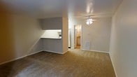 This single-floor unit features an open-concept layout that connects the main living area to the kitchen and bathroom with a functional, straightforward design. The living area is generously proportioned, finished with wall-to-wall carpeting in a neutral tan tone that adds warmth underfoot. A ceiling fan with an integrated light fixture is centrally mounted overhead, providing both air circulation and ambient lighting throughout the space.

A half-wall with a pass-through counter separates the kitchen from the living area, offering a degree of openness while maintaining a defined boundary between the two spaces. The kitchen is equipped with a full-size refrigerator and additional appliances, with under-cabinet lighting illuminating the workspace. The neutral gray wall tones throughout create a cohesive, understated palette.

A doorway from the main living area leads directly into a bathroom, which features tile flooring, a vanity with sink, and additional storage. A floor-level louvered vent is positioned along the wall, indicating a built-in HVAC system. White baseboards and trim throughout provide clean contrast against the gray walls, contributing to a tidy, well-maintained overall appearance.