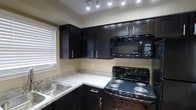 This single-floor space presents a thoughtfully designed interior centered around a modern, functional kitchen. The kitchen features rich, dark espresso-toned cabinetry that extends from base to upper levels, providing ample storage throughout. Light granite countertops offer a striking contrast against the dark cabinets, creating a visually balanced and contemporary aesthetic. The kitchen is equipped with black appliances, including an electric range with coil burners, an over-the-range microwave, and a stainless steel sink set beneath a window with white horizontal blinds that allow natural light to filter into the space. Recessed track lighting on the ceiling ensures bright, even illumination across the workspace. The layout follows an efficient L-shaped configuration, making practical use of the available space while maintaining an open and accessible flow. Neutral warm-toned walls complement the overall color palette, contributing to a clean, cohesive ambiance. The combination of dark cabinetry, light stone surfaces, and black appliances gives the kitchen a polished, modern character suited for everyday functionality and comfortable living.