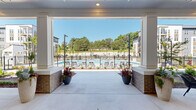 Located at 211 Hammond Center Drive in Raleigh, NC, this outdoor amenity space is designed to deliver a resort-like experience within a modern residential community. The centerpiece is a generously sized swimming pool enclosed by sleek black iron fencing, surrounded by an expansive concrete pool deck furnished with lounge chairs — ideal for relaxing under the Carolina sun. Mature trees and open green space beyond the fence provide a natural backdrop, enhancing the sense of seclusion and serenity.

A large covered pavilion anchors one end of the amenity area, featuring a coffered white ceiling, recessed lighting, and open-air sides that invite natural breezes while offering shade. This versatile space includes a ping-pong table, comfortable lounge seating with area rugs, and direct sightlines to the pool — making it equally suited for active recreation or casual socializing. Decorative planters with vibrant foliage add warmth and color throughout.

The surrounding grounds are thoughtfully landscaped with neatly trimmed shrubs, flowering plants, and mulched garden beds lining wide, well-lit walking paths that connect the amenity zone to the broader community. The multi-story residential buildings, finished in a contemporary palette of navy, gray, and warm brick, frame the space on multiple sides, creating a cohesive and inviting courtyard atmosphere.