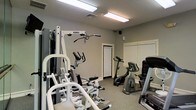 This fitness room is designed to support a well-rounded exercise routine in a dedicated, single-floor space. The room is equipped with a multi-function cable weight machine, offering a variety of strength-training exercises, and is complemented by multiple stationary bicycles — including an upright and a recumbent model — catering to a range of cardio preferences. Additional exercise equipment is positioned throughout the room, making efficient use of the available floor space. The interior features neutral gray walls, carpeted flooring, and a textured ceiling outfitted with fluorescent lighting that ensures the space is well-lit throughout. A window with horizontal blinds allows for natural light while maintaining privacy. Ventilation is provided via a ceiling vent, contributing to a comfortable workout environment. A standard interior door provides access to an adjoining area. The overall layout is practical and organized, offering users a straightforward and functional space for both cardiovascular and resistance training activities.