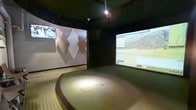 This dedicated golf simulator bay is thoughtfully designed to deliver an immersive, technology-driven experience in a compact, self-contained environment. The space centers on a raised, carpeted hitting platform with a clearly defined tee area, elevated slightly above the surrounding floor to replicate an authentic stance and swing setup. The dark ceiling and walls create a focused atmosphere, minimizing visual distractions and enhancing screen visibility.

A large projection screen dominates the front wall, displaying a detailed, photorealistic golf course simulation complete with a scoreboard and course statistics. A second flat-panel monitor is mounted to the left wall, offering supplemental viewing for replays or additional data. The side wall features a decorative geometric diamond-pattern panel in neutral tones, adding a refined aesthetic element that softens the otherwise dark, utilitarian finishes.

Overhead, recessed lighting provides targeted illumination directly over the hitting platform, ensuring excellent visibility at the tee. The overall design balances function and comfort, making the space well-suited for both casual rounds and serious practice sessions in a climate-controlled, year-round setting.