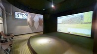 This dedicated golf simulator bay is thoughtfully designed to deliver an immersive, technology-driven experience in a compact, self-contained environment. The space centers on a raised, carpeted hitting platform with a clearly defined tee area, elevated slightly above the surrounding floor to replicate an authentic stance and swing setup. The dark ceiling and walls create a focused atmosphere, minimizing visual distractions and enhancing screen visibility.

A large projection screen dominates the front wall, displaying a detailed, photorealistic golf course simulation complete with a scoreboard and course statistics. A second flat-panel monitor is mounted to the left wall, offering supplemental viewing for replays or additional data. The side wall features a decorative geometric diamond-pattern panel in neutral tones, adding a refined aesthetic element that softens the otherwise dark, utilitarian finishes.

Overhead, recessed lighting provides targeted illumination directly over the hitting platform, ensuring excellent visibility at the tee. The overall design balances function and comfort, making the space well-suited for both casual rounds and serious practice sessions in a climate-controlled, year-round setting.